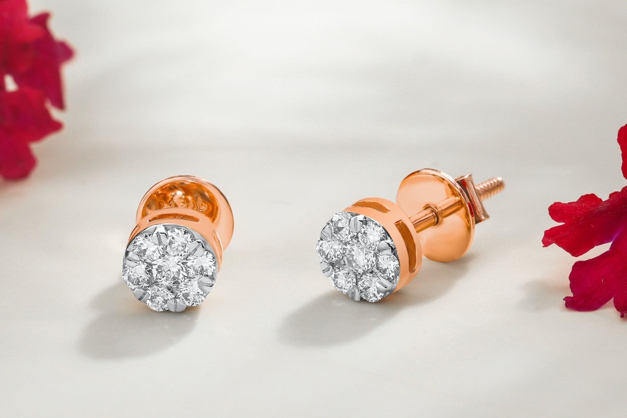 a pair of rose gold diamond stud earrings sit by red flower petals on a white fabric.